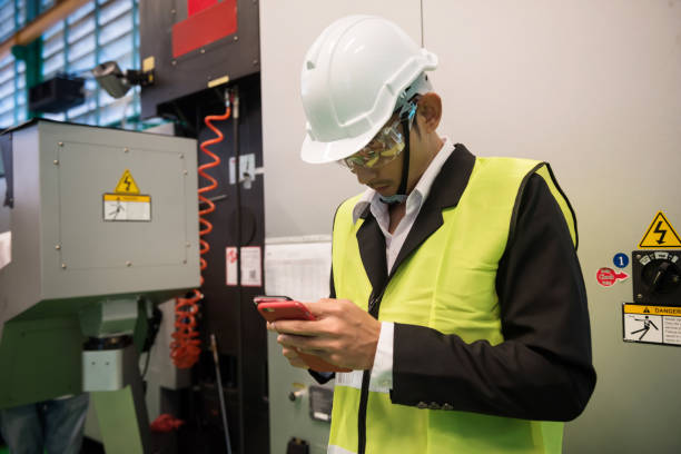 Busy factory Asian engineer manager checking manufacturing project plan from 2 smartphone devices near production machine with safety warning symbol.